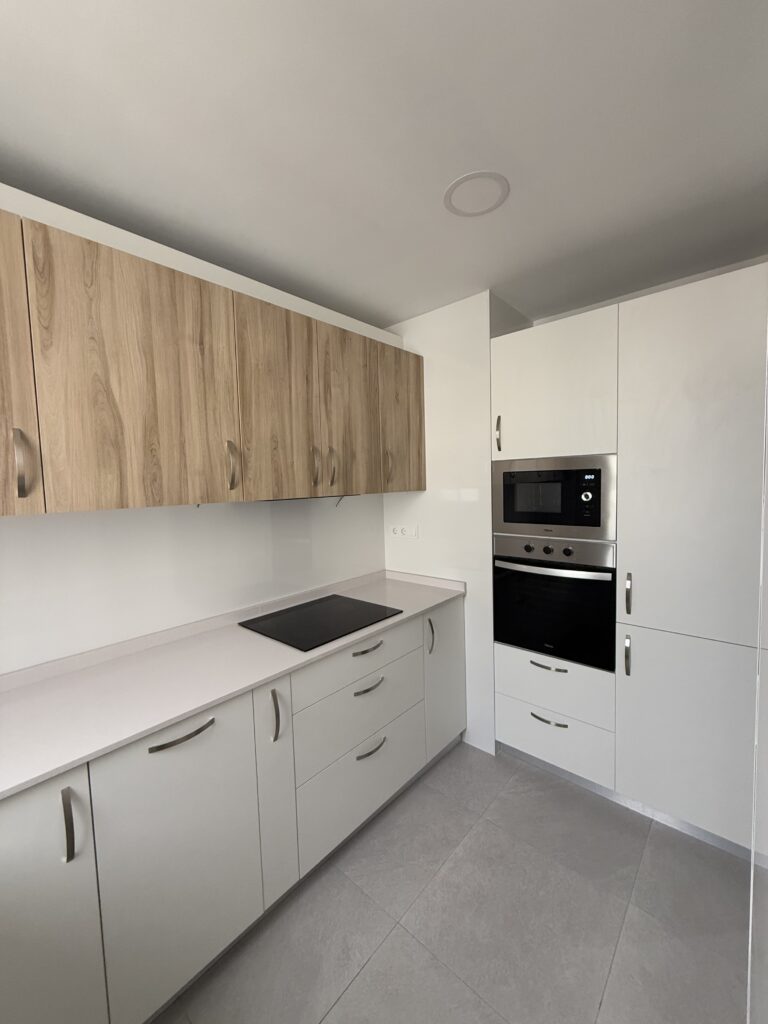 Esta cocina destaca por su diseño moderno y funcional, donde la elegancia se combina con la calidez de los acabados naturales. Los muebles inferiores en blanco mate aportan una sensación de amplitud y luminosidad, mientras que los armarios superiores con textura de madera clara añaden un toque acogedor y natural al espacio. La encimera continua de tono claro resalta la estética y limpieza. Es una cocina perfecta para disfrutar tanto de la funcionalidad como del diseño.

